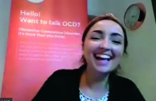 Picture of a smiling Zoe sitting in front of an orange OCD-UK banner with the words 'Hello, want to talk OCD?'